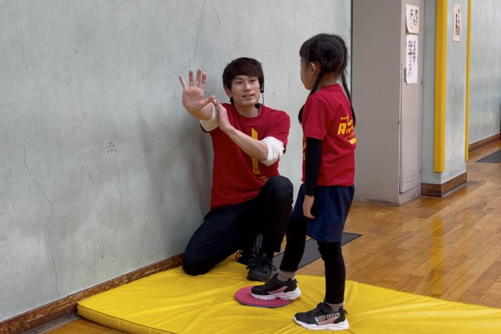 野々市教室にて、体操競技における壁倒立の「腕を伸ばすコツ」を指導する笑顔の大久保先生と、話を聞く幼児の女児