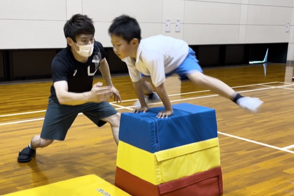加賀市教室にて、カッコよく跳び箱の縦を跳ぶ小学生の男児の子供と、指導する大久保先生