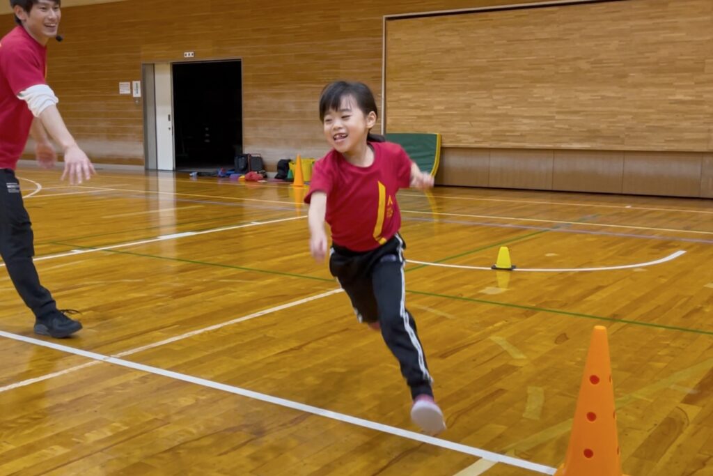福井市の体操教室「大久保先生のスポーツレベルアップアカデミー」にて、オリジナルの切り返しトレーニング「先生をよけろ」を行う、満面の笑顔の小学生女児と笑顔で見守る大久保先生