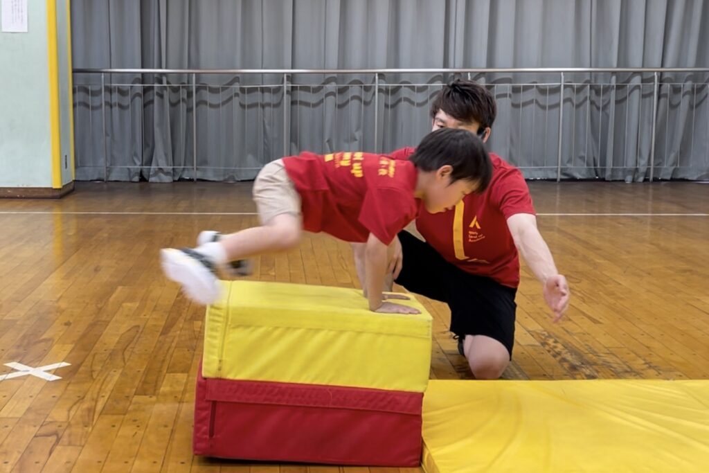 野々市教室にて、跳び箱の縦をかっこいいフォームでとぶ小学生の子供とサポートをする大久保先生