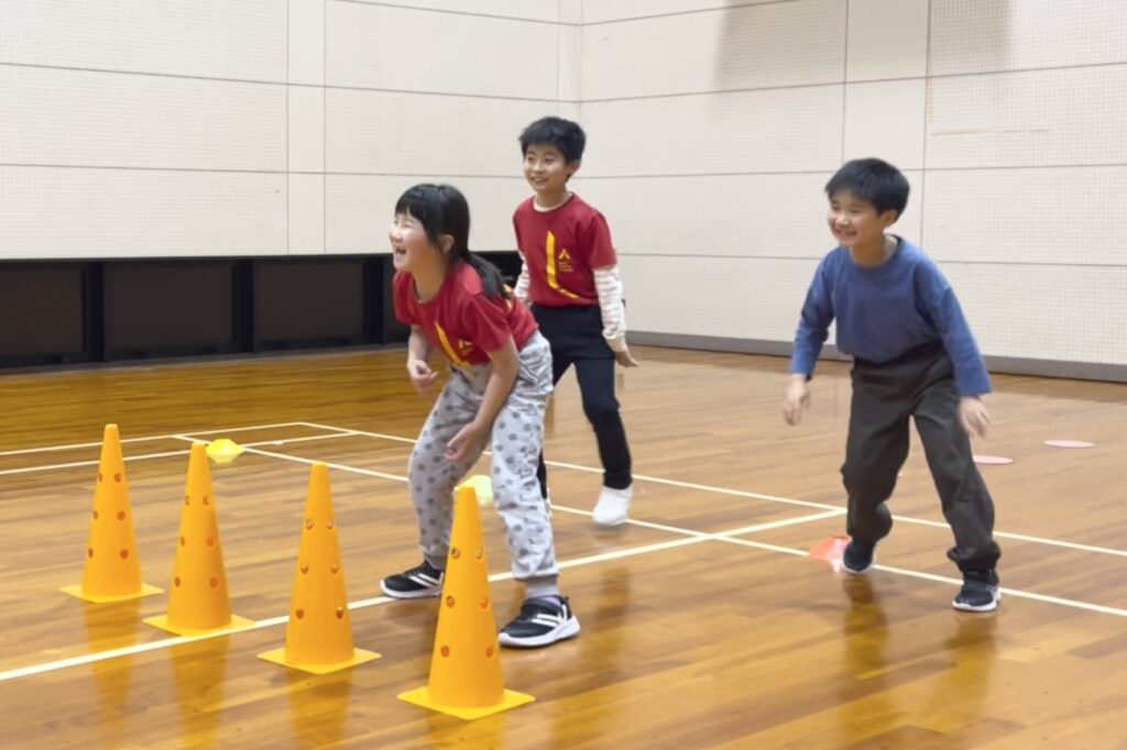 加賀市の体操教室「大久保先生のスポーツレベルアップアカデミー加賀市教室」にて、オリジナル運動プログラム「壁鬼ごっこ」を楽しむ幼児と小学生の子どもたち