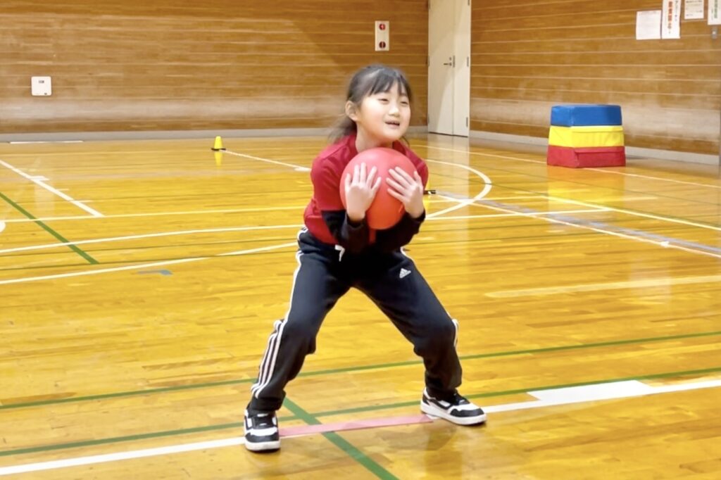 福井市の体操教室「大久保先生のスポーツレベルアップアカデミー福井市教室」にて、ドッジボールのキャッチ姿勢を練習する笑顔の女児|下から包み込む取り方