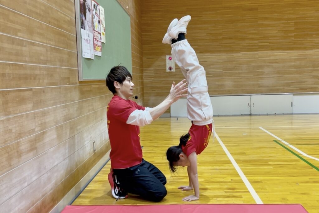体操教室「大久保先生のスポーツレベルアップアカデミー福井市教室」にて、小学生が補助なし倒立にチャレンジし大久保先生が安全にサポートする様子