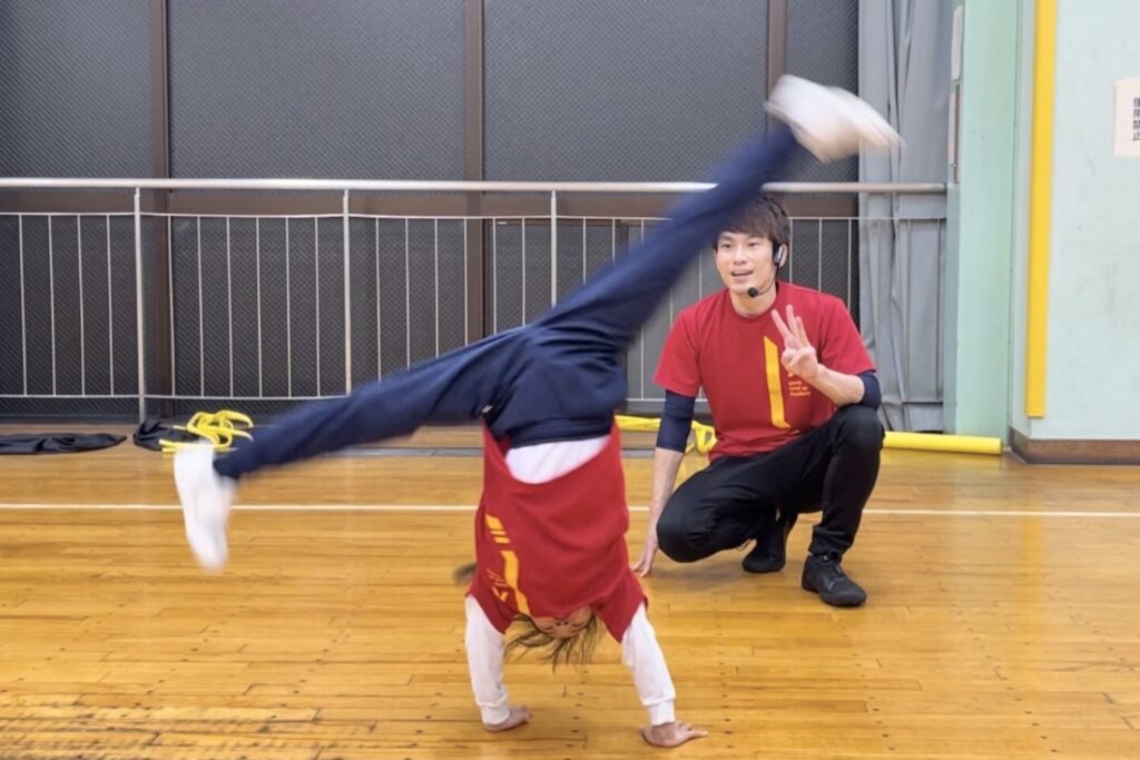 野々市の体操教室「大久保スポーツ教室野々市会場」にて、子どもが体操競技の技「側転（側方倒立回転）」をかっこいいフォームで行い、大久保先生が笑顔で見守っている様子