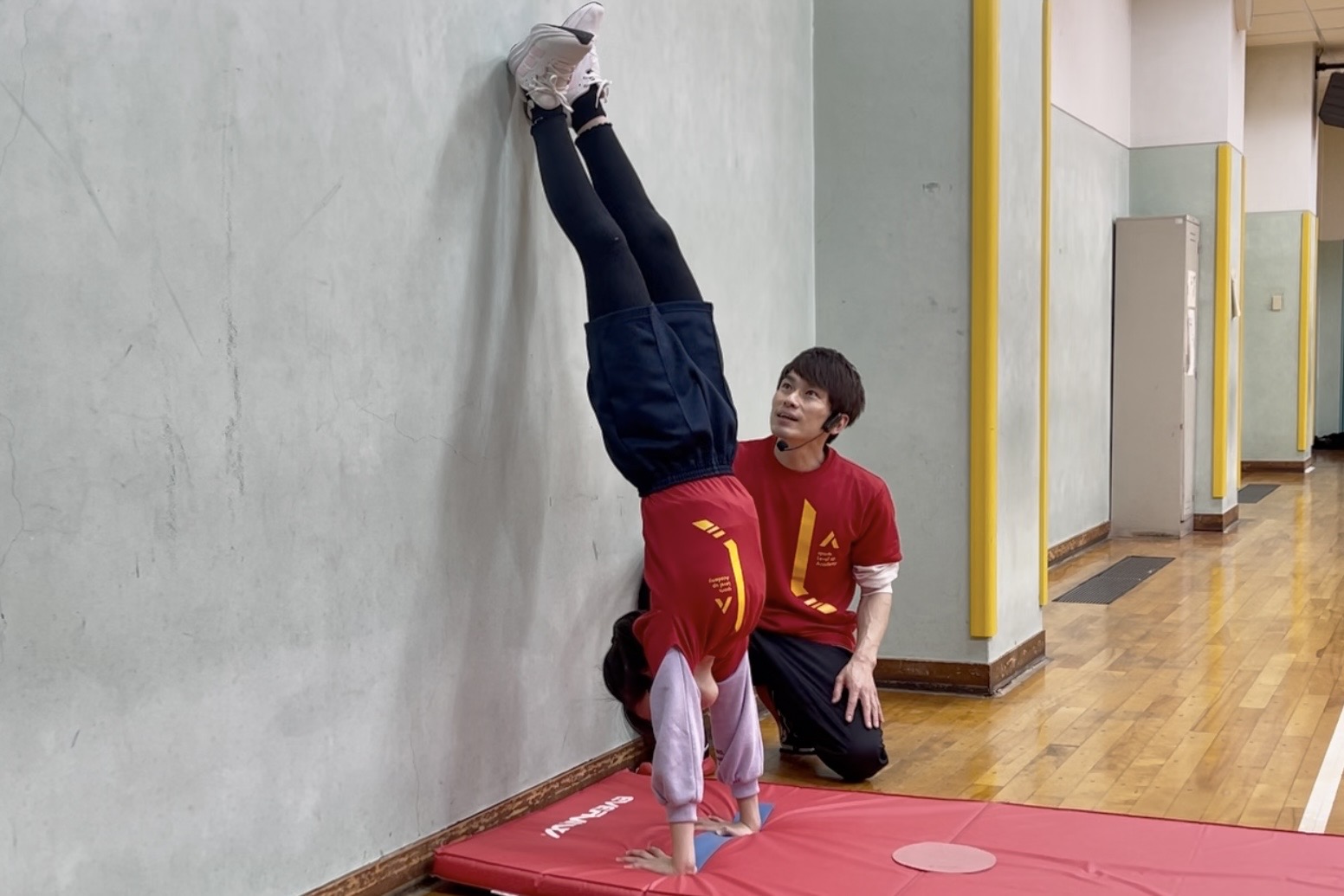 野々市の体操教室で、壁倒立をかっこよく行う子供を大久保先生が笑顔で見守っている様子