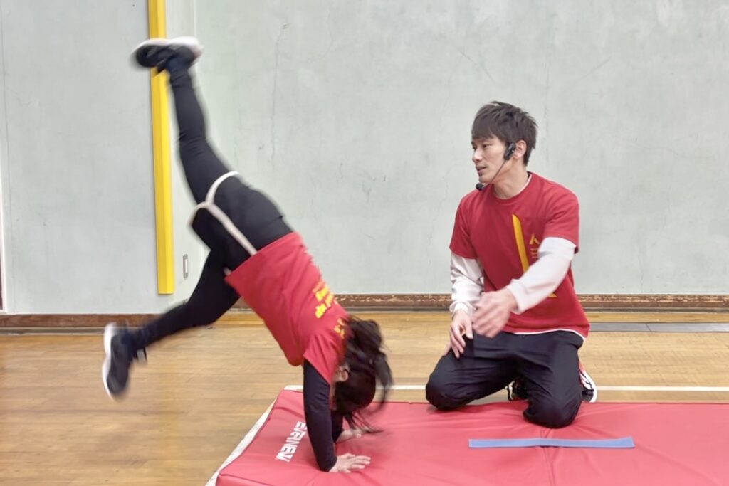 野々市教室の体操競技のマット運動の単元にて「倒立」に挑戦する幼児と、サポート姿勢に入る大久保先生