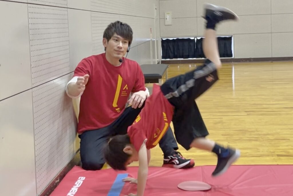 小松市教室にて、マット運動の「壁倒立」にチャレンジする子供と、笑顔で見守る大久保先生。