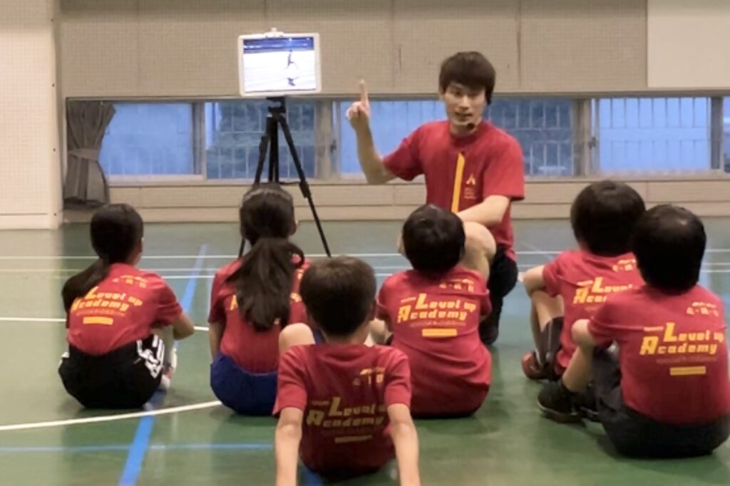 白山市会場にて、タブレットを用いて運動のポイントを子供達に解説する大久保先生と、座ってその説明を真剣に聞く子供達