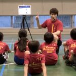 白山市会場にて、タブレットを用いて運動のポイントを子供達に解説する大久保先生と、座ってその説明を真剣に聞く子供達